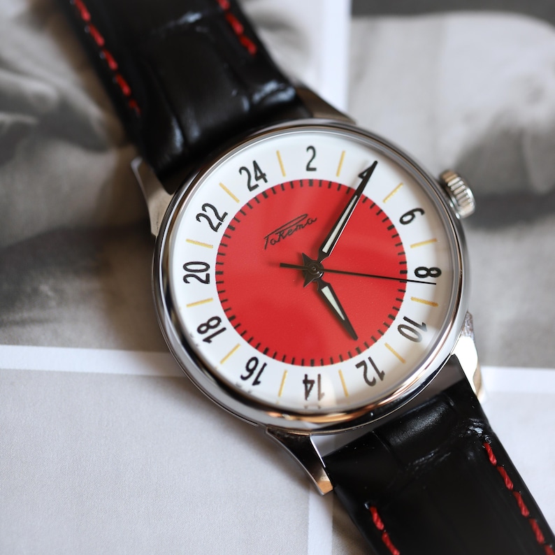 May include: A wristwatch with a red and white face, black hands, and a black leather band. The watch face features the brand name "Raketa" and a 24-hour clock display. The band has red stitching.