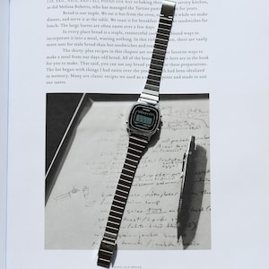 K&ouml;nnte beinhalten: Eine silberfarbene Digitalarmbanduhr mit rechteckigem Zifferblatt und flexiblem Metallgliederarmband. Die Uhr hat einen schwarzen Displayrand mit dem Casio-Markennamen und dem in Wei&szlig; gedruckten Text Water Resist. Ein d&uuml;nner Metallstift liegt daneben.