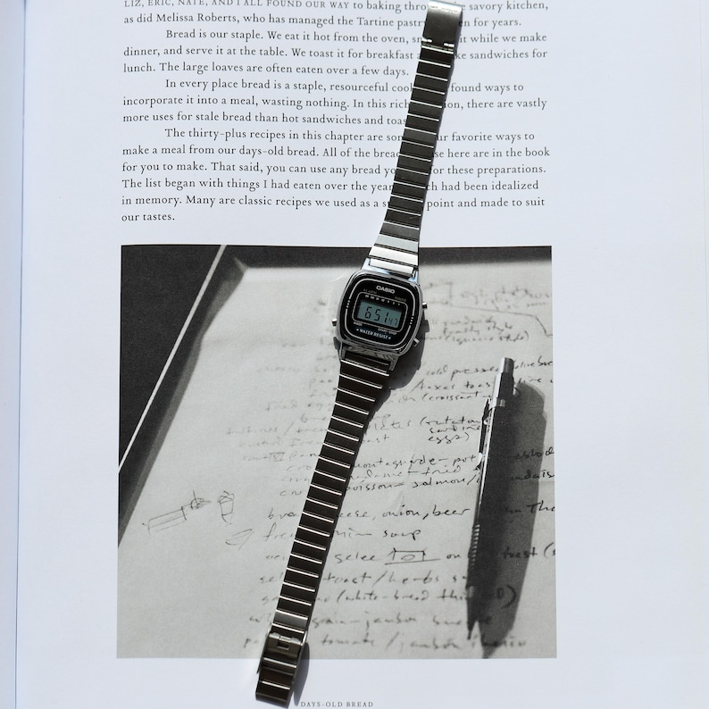 K&ouml;nnte beinhalten: Eine silberfarbene Digitalarmbanduhr mit rechteckigem Zifferblatt und flexiblem Metallgliederarmband. Die Uhr hat einen schwarzen Displayrand mit dem Casio-Markennamen und dem in Wei&szlig; gedruckten Text Water Resist. Ein d&uuml;nner Metallstift liegt daneben.