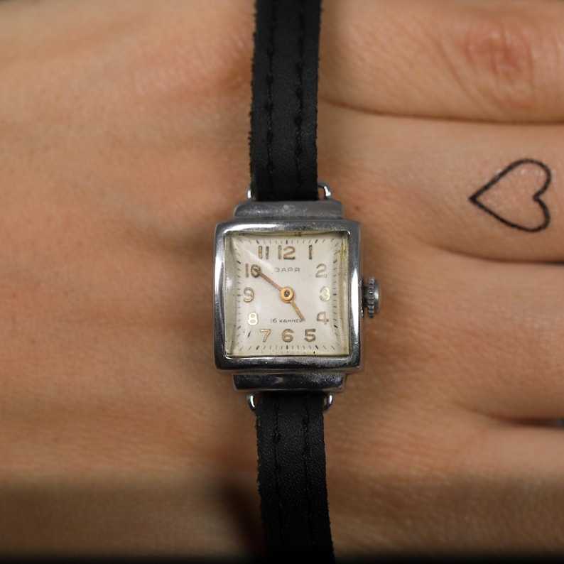 May include: A silver square-shaped wristwatch with a white face and black leather band. The watch face has black Roman numeral hour markers and a small second hand. The watch face also has the text "ЗАРЯ 15 камней" printed on it.