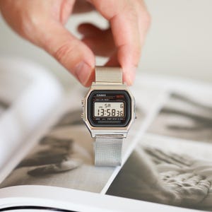 Puede incluir: Se muestra un reloj digital plateado con una correa de malla. La esfera del reloj muestra la hora 13:58:36. El reloj tiene una esfera rectangular con detalles en blanco y negro. El reloj se sostiene sobre una superficie con estampado en blanco y negro.