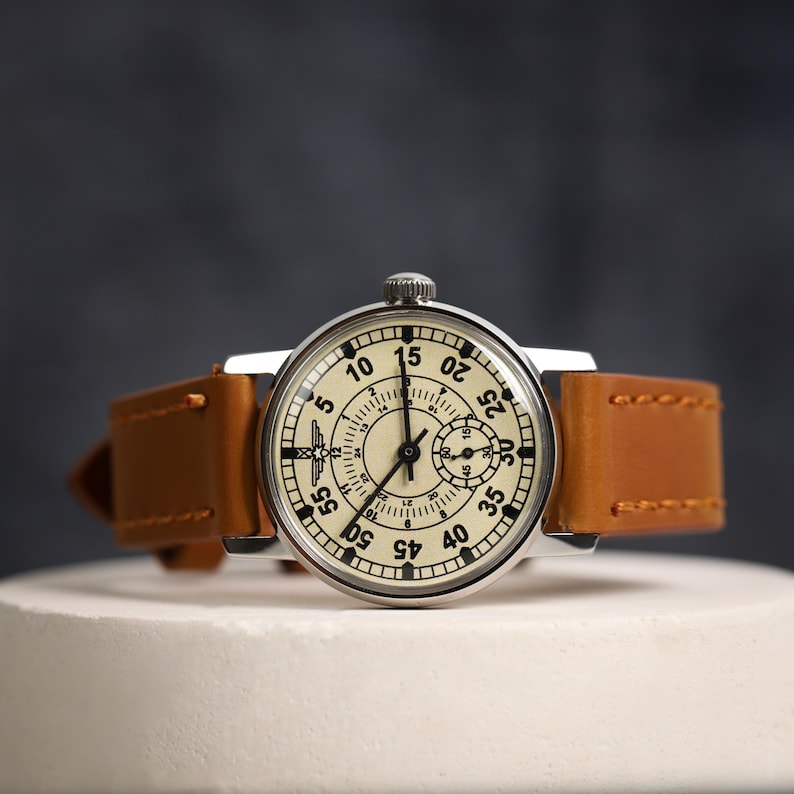 May include: A silver wristwatch with a white face and black markings. The watch has a brown leather band with white stitching. The face of the watch has numbers from 5 to 55 and a smaller circle in the center with numbers from 1 to 12.