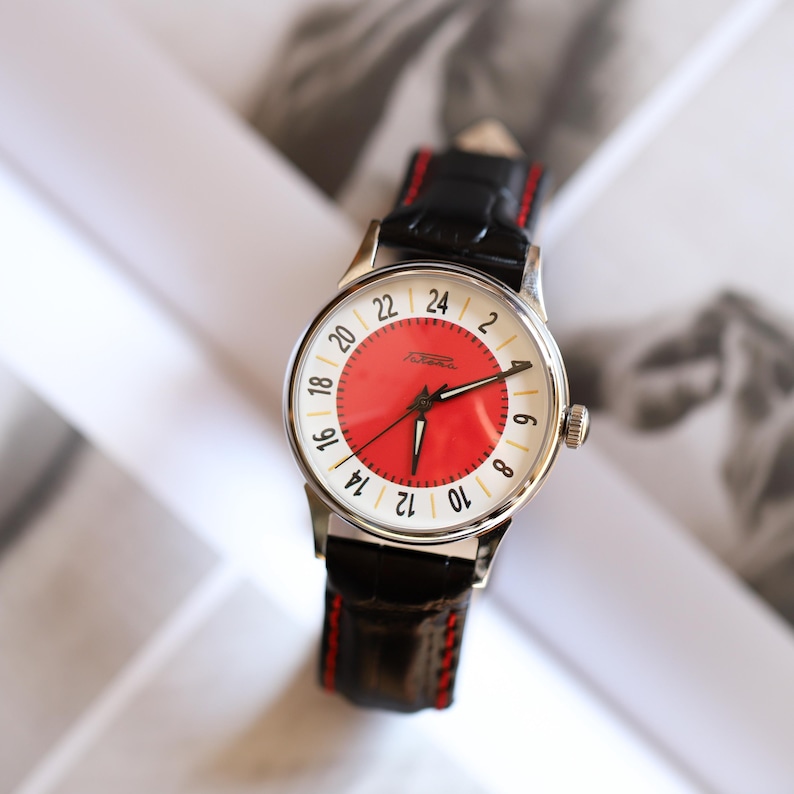 May include: A vintage wristwatch with a silver case, a red and white dial, and a black leather band with red stitching. The watch face displays the numbers 2 to 24. The watch has a classic, retro design.