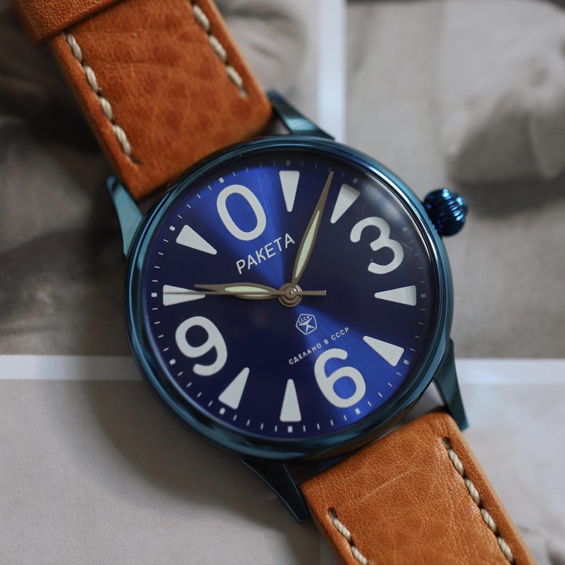 May include: A blue Paketa wristwatch with a tan leather strap. The watch face features large white numerals and hands, with the brand name "Paketa". The strap is stitched with white thread.