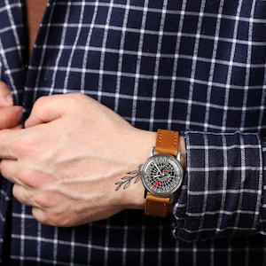 May include: A man wearing a blue and white plaid shirt with a brown leather wristwatch with a black face. The watch has a unique design with a compass-like face.