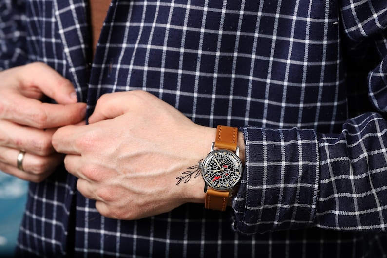 May include: A man wearing a blue and white plaid shirt with a brown leather wristwatch with a black face. The watch has a unique design with a compass-like face.