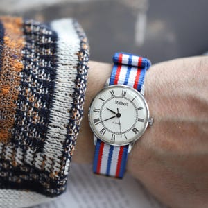 Könnte beinhalten: Eine Sekonda-Armbanduhr mit weißem Zifferblatt, schwarzen Zeigern und römischen Ziffern. Die Uhr hat ein silbernes Gehäuse und ein blau-weiß-rot gestreiftes Nylonarmband. Die Uhr befindet sich an einem Handgelenk.