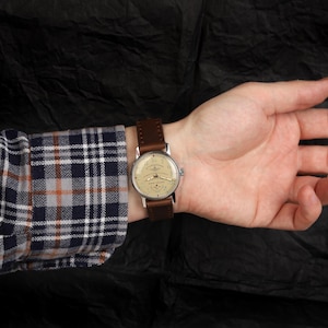 Pode incluir: Um rel&oacute;gio de pulso com uma pulseira de couro marrom e um mostrador prateado. O rel&oacute;gio tem um mostrador branco com marcas pretas e uma janela de data na posi&ccedil;&atilde;o das 3 horas.