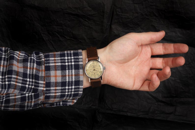 Pode incluir: Um rel&oacute;gio de pulso com uma pulseira de couro marrom e um mostrador prateado. O rel&oacute;gio tem um mostrador branco com marcas pretas e uma janela de data na posi&ccedil;&atilde;o das 3 horas.
