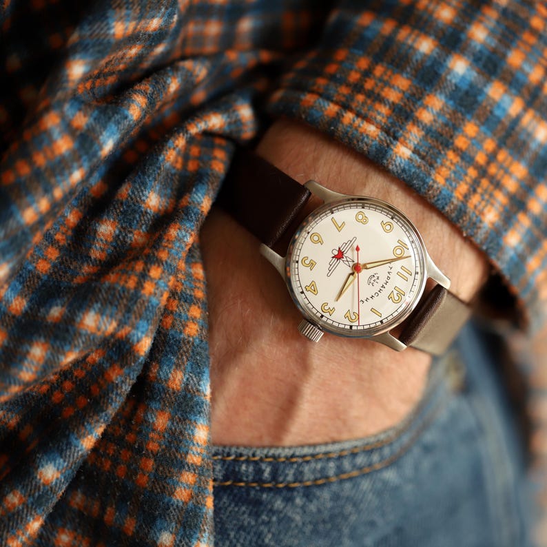 May include: A vintage wristwatch with a cream-colored face, featuring gold numerals and hands, and a red emblem. The watch has a silver case and a dark brown leather band. The watch is worn on a wrist, with a blue and orange plaid shirt in the background.