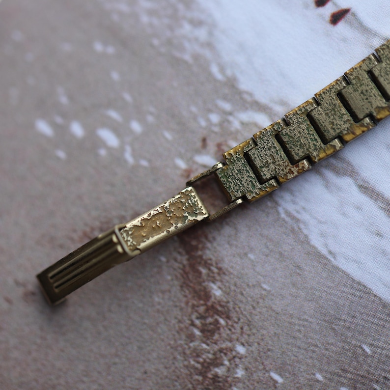 Puede incluir: Primer plano de una correa de reloj dorada vintage. La correa presenta un dise&ntilde;o de eslabones rectangulares con un aspecto texturizado y envejecido. El cierre es visible, mostrando signos de desgaste y p&aacute;tina, lo que sugiere su antig&uuml;edad.