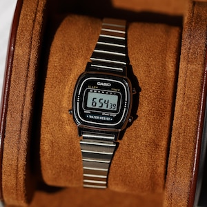 K&ouml;nnte beinhalten: Eine silberfarbene Digitalarmbanduhr mit Metallgliederarmband und schwarzem rechteckigem Zifferblatt. Das Display zeigt die Uhrzeit 6:54:39, mit dem in Wei&szlig; aufgedruckten Text CASIO, ALARM, TIMER, MODE, START-STOP und WATER RESIST.