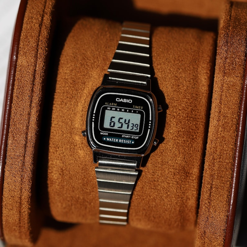 K&ouml;nnte beinhalten: Eine silberfarbene Digitalarmbanduhr mit Metallgliederarmband und schwarzem rechteckigem Zifferblatt. Das Display zeigt die Uhrzeit 6:54:39, mit dem in Wei&szlig; aufgedruckten Text CASIO, ALARM, TIMER, MODE, START-STOP und WATER RESIST.