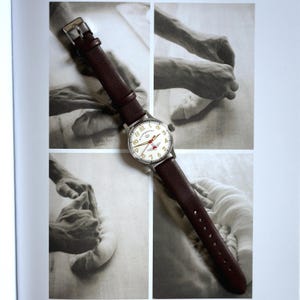Könnte beinhalten: Eine Armbanduhr mit silberfarbenem Gehäuse und cremefarbenem Zifferblatt. Sie hat schwarze Ziffern und rote Akzente. Das Armband ist aus braunem Leder. Die Uhr ist vor einem Hintergrund aus Schwarz-Weiß-Bildern platziert.
