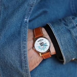 Puede incluir: Un reloj de pulsera dorado con una esfera blanca que presenta un gr&aacute;fico en blanco y negro y detalles en azul. Tiene una correa de cuero marr&oacute;n. El reloj se lleva en la mu&ntilde;eca, con una chaqueta vaquera azul de fondo.