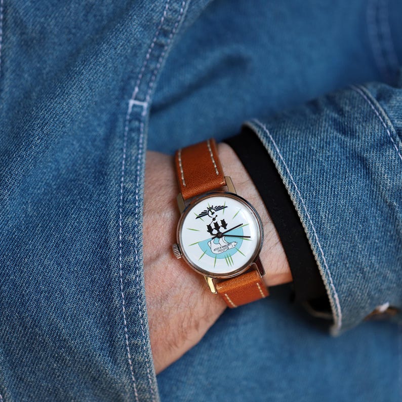 Puede incluir: Un reloj de pulsera dorado con una esfera blanca que presenta un gr&aacute;fico en blanco y negro y detalles en azul. Tiene una correa de cuero marr&oacute;n. El reloj se lleva en la mu&ntilde;eca, con una chaqueta vaquera azul de fondo.