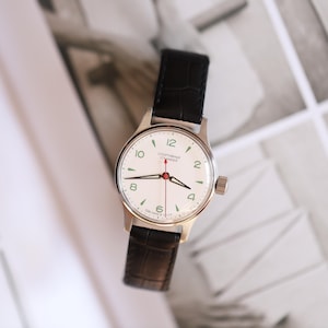 May include: A vintage wristwatch with a white face, black leather band, and silver-colored case. The watch face features black numerals, green accents, and red hands. The watch is a classic timepiece.