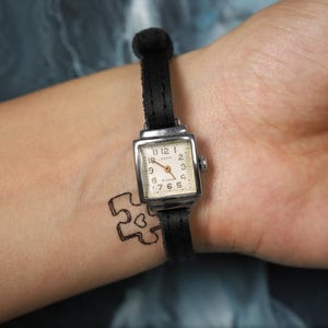 May include: A black leather wristwatch with a silver square face and white clock hands. The watch face has black Roman numeral markings and the text "12, 1, 2, 3, 4, 5, 6, 7, 8, 9, 10, 11" around the edge. The watch is on a person's wrist.