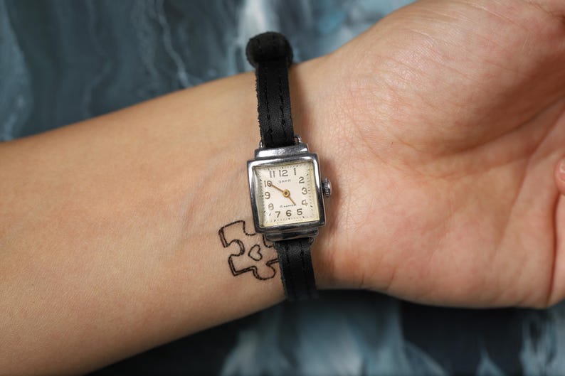 May include: A black leather wristwatch with a silver square face and white clock hands. The watch face has black Roman numeral markings and the text "12, 1, 2, 3, 4, 5, 6, 7, 8, 9, 10, 11" around the edge. The watch is on a person's wrist.