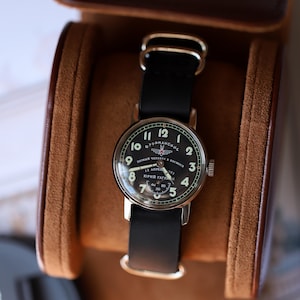 Könnte beinhalten: Eine schwarze Armbanduhr mit schwarzem Lederarmband in einem braunen Lederetui. Das schwarze Zifferblatt zeigt weiße Ziffern und Text, darunter "Первый человек в космосе" und "Юрий Гагарин".
