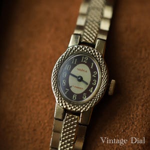Puede incluir: Un reloj de pulsera vintage dorado con esfera marr&oacute;n oscuro y n&uacute;meros dorados. La esfera del reloj tiene la palabra cir&iacute;lica "ЧАЙКА" y "17 KAMHEЙ". La correa del reloj tiene un dise&ntilde;o texturizado. Las palabras "Vintage Dial" est&aacute;n en la esquina inferior derecha.