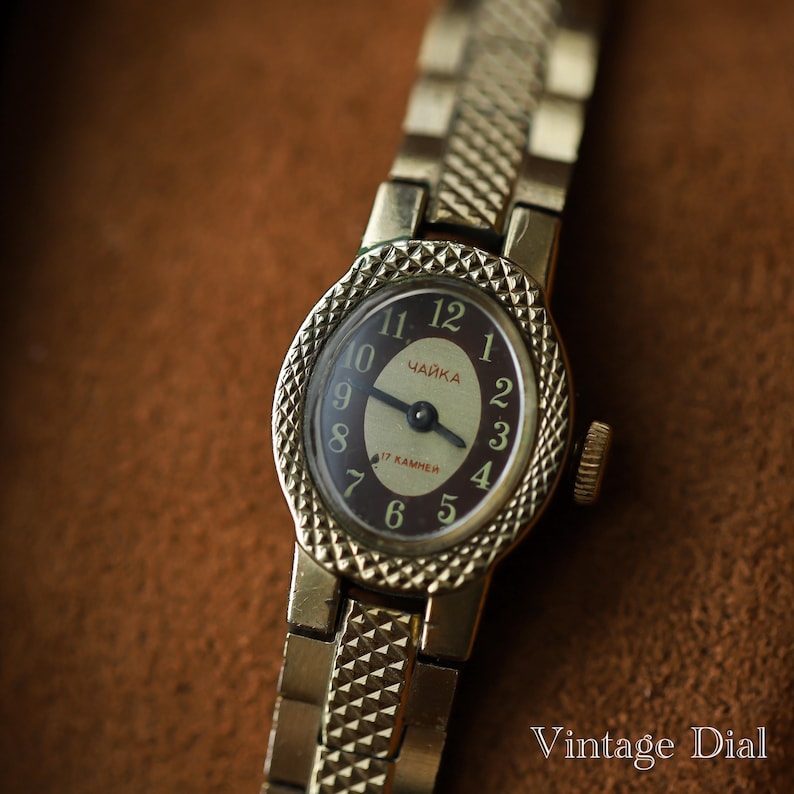 Puede incluir: Un reloj de pulsera vintage dorado con esfera marr&oacute;n oscuro y n&uacute;meros dorados. La esfera del reloj tiene la palabra cir&iacute;lica "ЧАЙКА" y "17 KAMHEЙ". La correa del reloj tiene un dise&ntilde;o texturizado. Las palabras "Vintage Dial" est&aacute;n en la esquina inferior derecha.