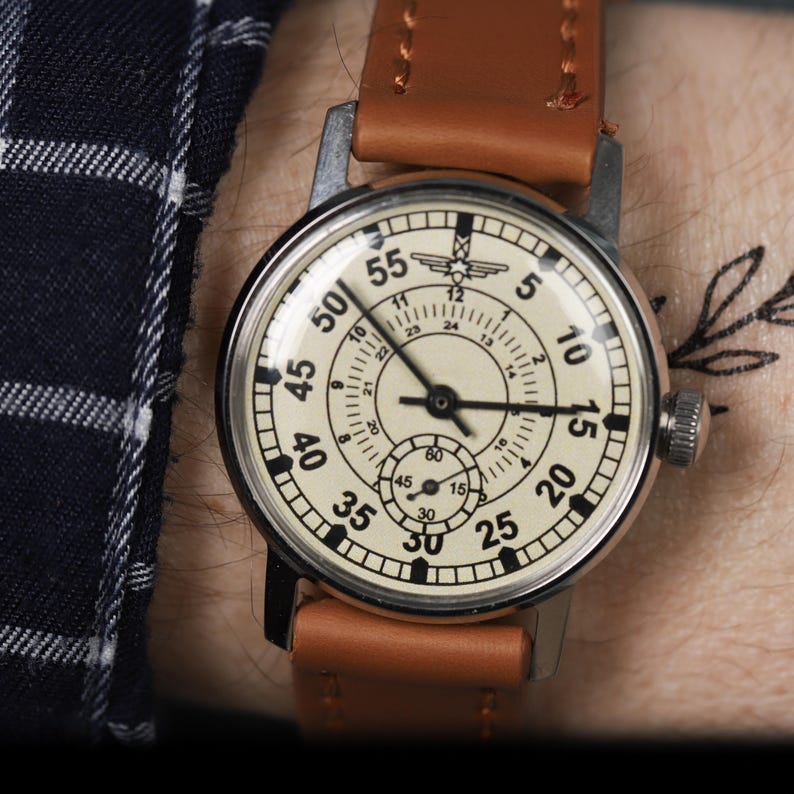 May include: A vintage wristwatch with a white face and black markings. The watch has a brown leather band and a silver case. The face of the watch has numbers from 5 to 55, and the words "60", "45", "30", and "15" are printed around the edge.