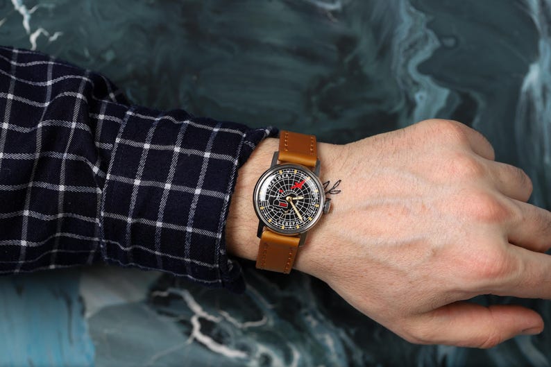 May include: A wristwatch with a brown leather band and a black face with white markings. The watch face has a red accent and a small red hand.