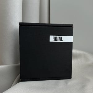 Puede incluir: Una caja cuadrada negra con una etiqueta blanca que dice "VINTAGE DIAL". La caja est&aacute; sobre una superficie de color claro. La caja est&aacute; cerrada y tiene un dise&ntilde;o simple y minimalista.