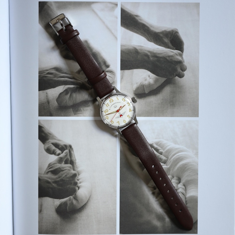 May include: A vintage wristwatch with a brown leather band and a silver-colored case. The watch face is cream-colored with black numerals and red accents. The watch is displayed on a page with black and white photographs.