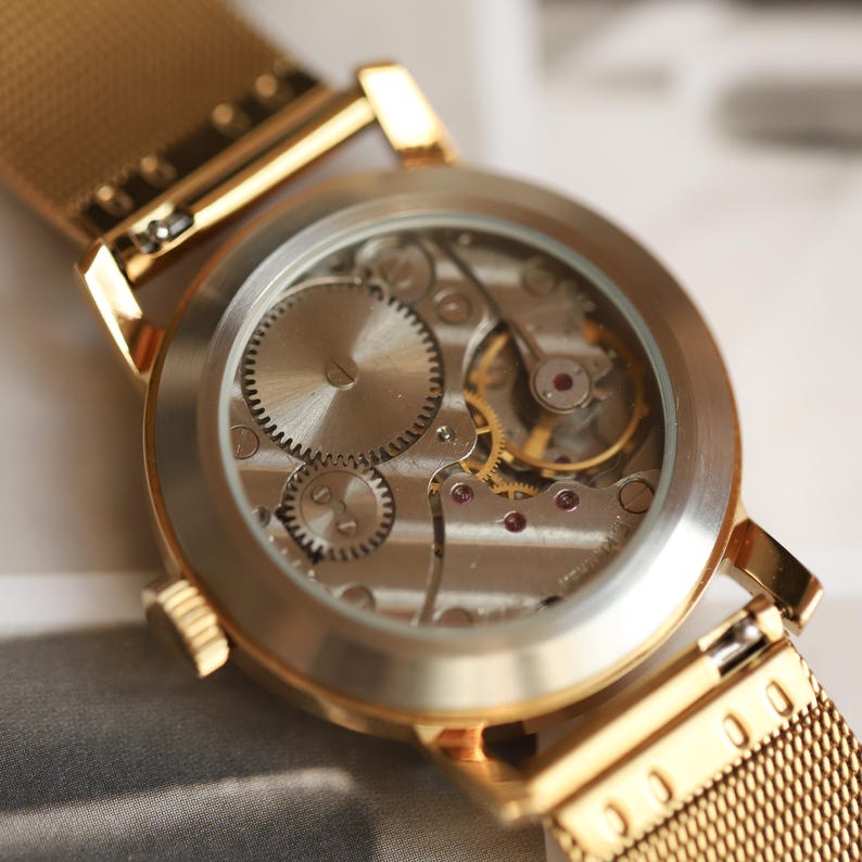 K&ouml;nnte beinhalten: Eine goldfarbene Armbanduhr mit transparentem Geh&auml;useboden, der das komplexe mechanische Uhrwerk mit sichtbaren Zahnr&auml;dern und kleinen Rubinlagern freigibt. Die Uhr ist mit einem feinen goldfarbenen Metall-Mesh-Armband ausgestattet.