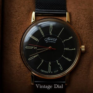 May include: A vintage wristwatch with a black dial and gold-colored case. The watch face features gold-colored hour markers and hands, with the text "Дымпел" and "23 KAMHA". The watch has a black band and the text "Vintage Dial" is at the bottom.