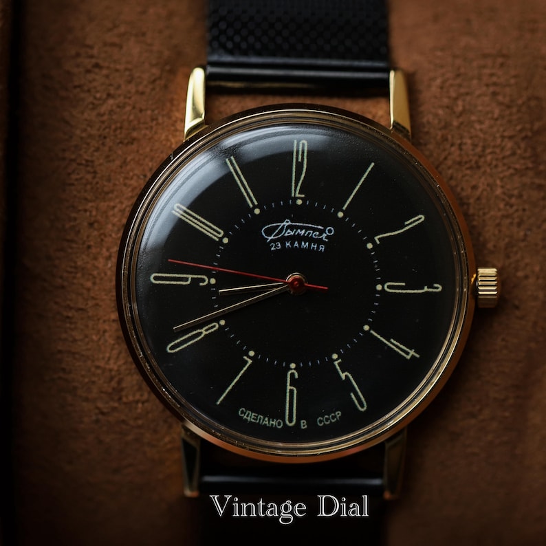 May include: A vintage wristwatch with a black dial and gold-colored case. The watch face features gold-colored hour markers and hands, with the text "Дымпел" and "23 KAMHA". The watch has a black band and the text "Vintage Dial" is at the bottom.