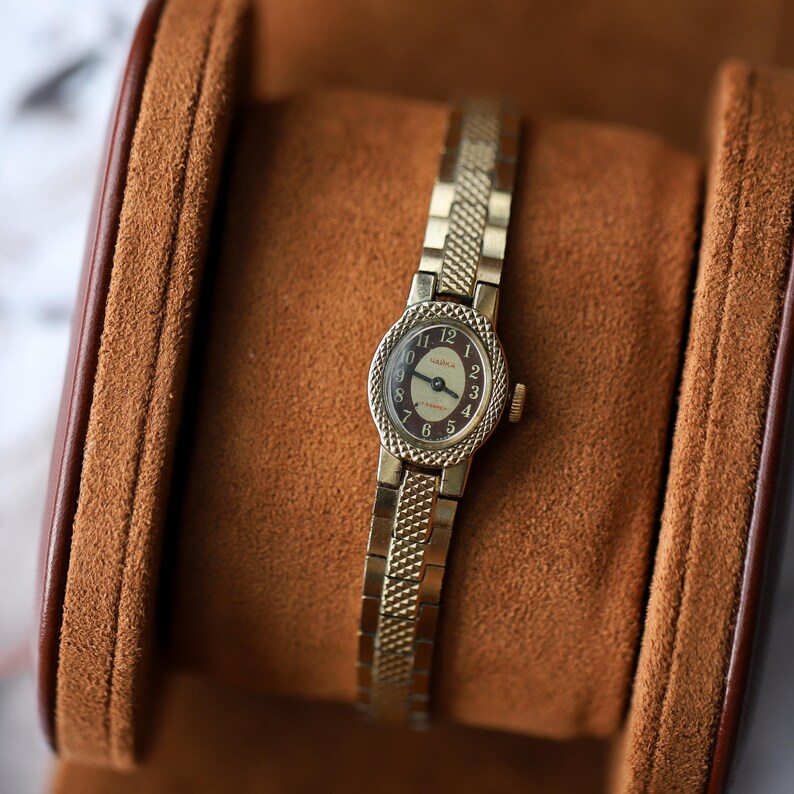 Puede incluir: Un reloj de pulsera vintage dorado con una correa texturizada y una peque&ntilde;a esfera ovalada. La esfera del reloj tiene un fondo claro con n&uacute;meros y agujas negras. El reloj se exhibe en un estuche forrado de cuero marr&oacute;n.