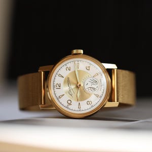 K&ouml;nnte beinhalten: Eine goldfarbene Armbanduhr mit rundem Geh&auml;use und passendem Mesh-Armband. Das wei&szlig;e Zifferblatt zeigt goldene arabische Ziffern, einen goldenen Mittelkreis und ein Hilfszifferblatt mit einer Grafik eines Satelliten im Erdorbit sowie kyrillischem Text.