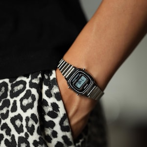 K&ouml;nnte beinhalten: Eine silberfarbene Digitalarmbanduhr mit einem Metallgliederarmband und einem rechteckigen schwarzen Zifferblatt. Die Uhr verf&uuml;gt &uuml;ber eine Digitalanzeige mit Uhrzeit sowie den Markennamen Casio und den Schriftzug Water Resist.