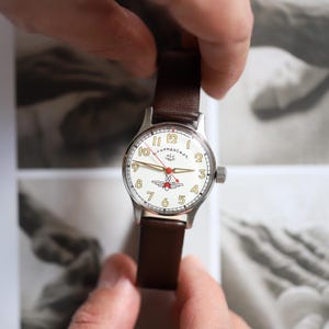 May include: A silver wristwatch with a white face, featuring black numerals and a red second hand. The watch has a brown leather band. The watch face includes the text "ТУРБАНСКИЕ".