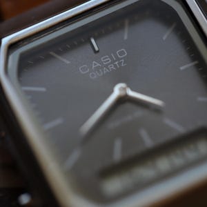 May include: Close-up of a Casio Quartz watch. The watch has a dark gray face with silver hands and hour markers. The brand name "CASIO QUARTZ" is printed in white. The watch has a rectangular shape with a silver-colored case.