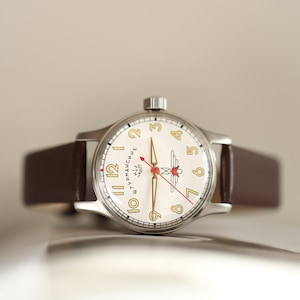 May include: A vintage-style wristwatch with a cream-colored face, gold numerals, and a red second hand. The watch has a silver case and a brown leather strap. The watch face features a logo and text. The watch is a classic timepiece.