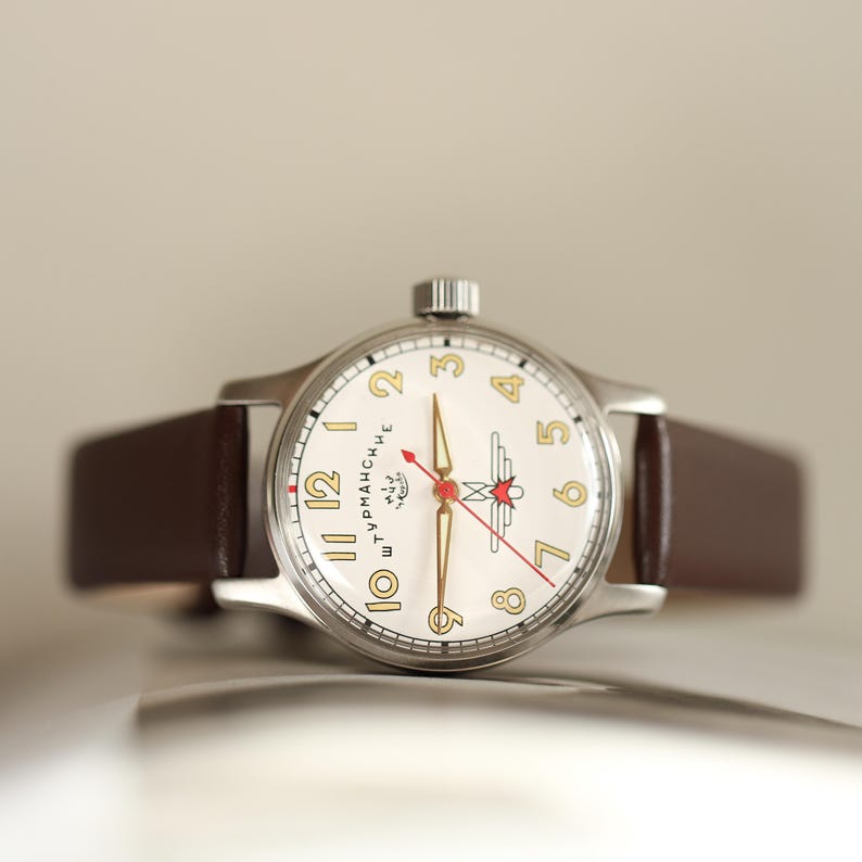 May include: A vintage-style wristwatch with a cream-colored face, gold numerals, and a red second hand. The watch has a silver case and a brown leather strap. The watch face features a logo and text. The watch is a classic timepiece.