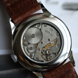 May include: Close-up of a vintage wristwatch's exposed movement. The watch features a silver-toned case, a brown leather strap, and intricate gears and springs. The movement is marked with the text "2809.H4".