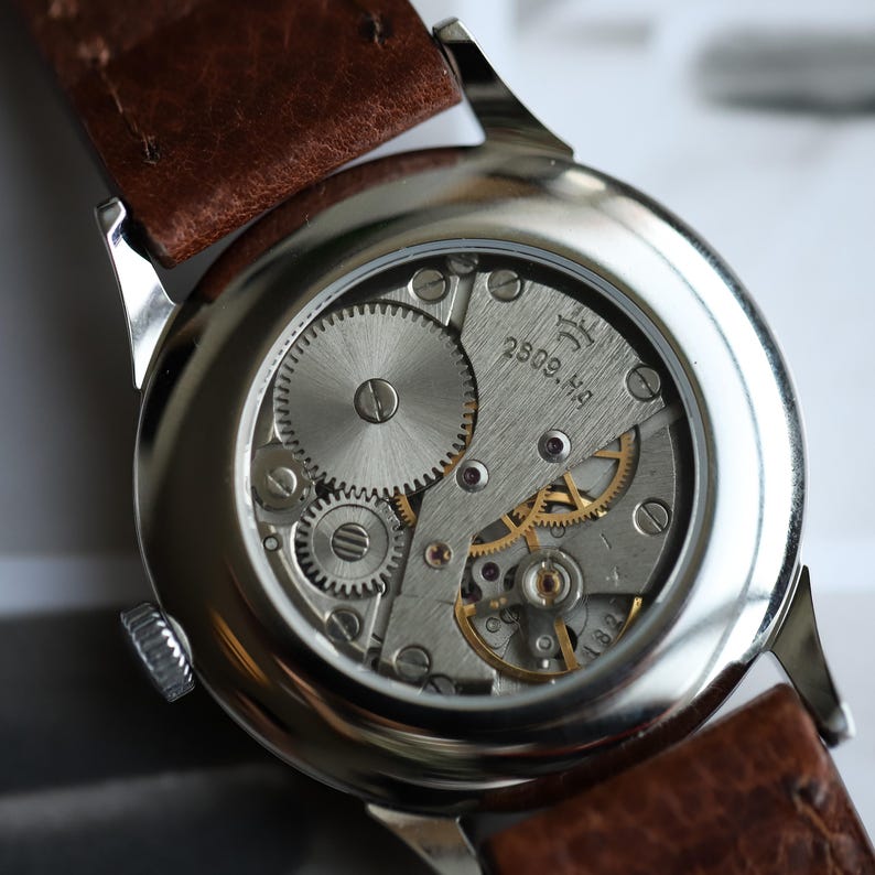 May include: Close-up of a vintage wristwatch's exposed movement. The watch features a silver-toned case, a brown leather strap, and intricate gears and springs. The movement is marked with the text "2809.H4".