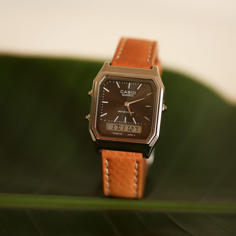 May include: A Casio Quartz watch with a brown leather band. The watch has a dark face with silver-tone accents and displays the time as 17:11:27. The watch is water resistant.