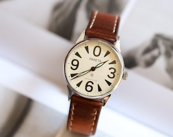 Könnte beinhalten: Eine Vintage-Armbanduhr mit cremefarbenem Zifferblatt, schwarzen Ziffern und dem Namen "PAKETA" auf dem Zifferblatt. Sie hat ein braunes Lederarmband mit weißen Nähten. Das Uhrengehäuse ist silberfarben.