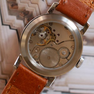 Puede incluir: Primer plano del mecanismo de un reloj de pulsera vintage, visible a trav&eacute;s de la parte trasera transparente. El reloj tiene una caja plateada y una correa de cuero marr&oacute;n. Los engranajes y joyas internos son visibles, destacando el intrincado dise&ntilde;o del reloj.