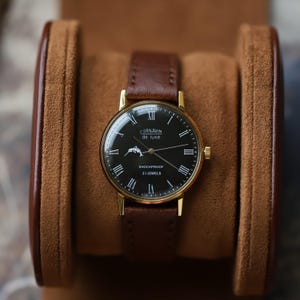 May include: A gold-tone wristwatch with a round black dial, featuring white Roman numeral hour markers, a dolphin graphic, and the text CORNAVIN de luxe SHOCKPROOF 23 JEWELS. The watch has a brown leather strap and a gold-tone metal case.