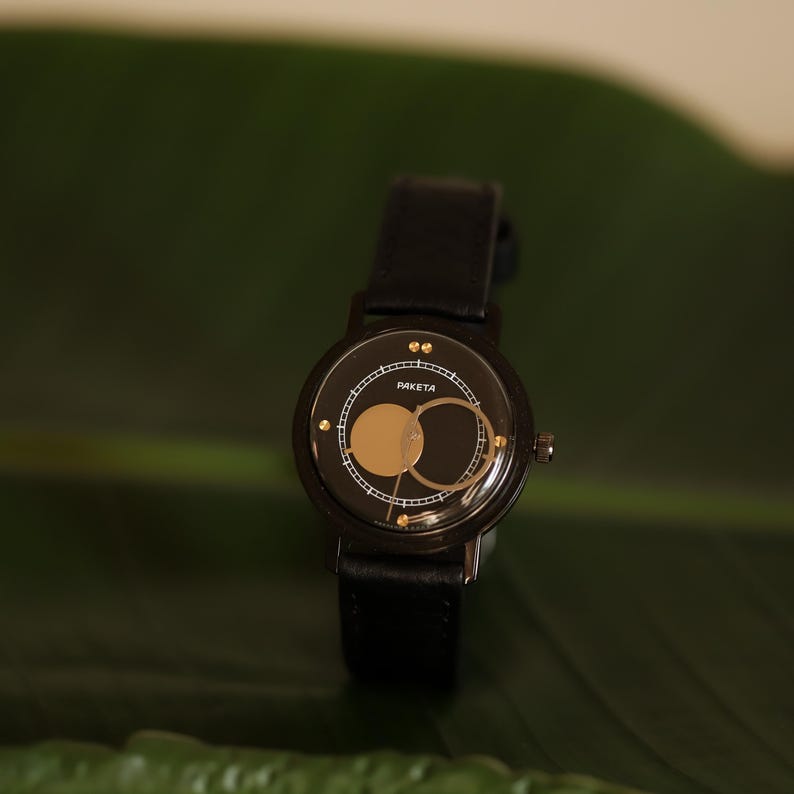 May include: Black wristwatch with a leather band. The watch face features a gold-colored moon phase design and the word "PAKETA" in white. The watch is set against a green leaf.
