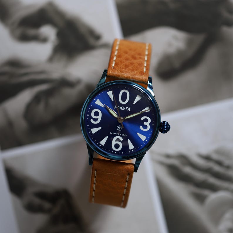 May include: A blue-faced wristwatch with white numerals and hands, and a brown leather strap. The watch face has the word "PAKETA" and Cyrillic text. The watch case is a dark blue colour. The watch strap is light brown with white stitching.