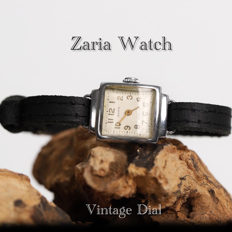 May include: A silver square-shaped wristwatch with a black leather band. The watch face is white with black Roman numeral hour markers and a small second hand. The watch face also has the text "ZAPCH" in black. The watch is sitting on a piece of wood. The text "Zaria Watch" is above the watch and the text "Vintage Dial" is below the watch.