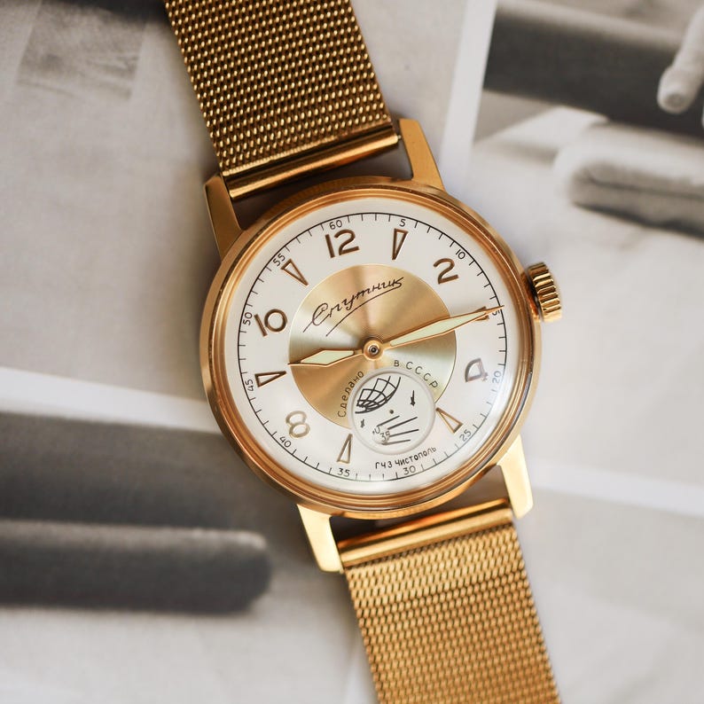 K&ouml;nnte beinhalten: Eine goldfarbene Armbanduhr mit Mesh-Armband und rundem wei&szlig;em Zifferblatt. Das Zifferblatt zeigt goldfarbene arabische Ziffern, einen zentralen goldfarbenen Akzent und kyrillischen Text, einschlie&szlig;lich des Wortes Sputnik. Ein kleines Hilfszifferblatt zeigt eine Weltkugel.